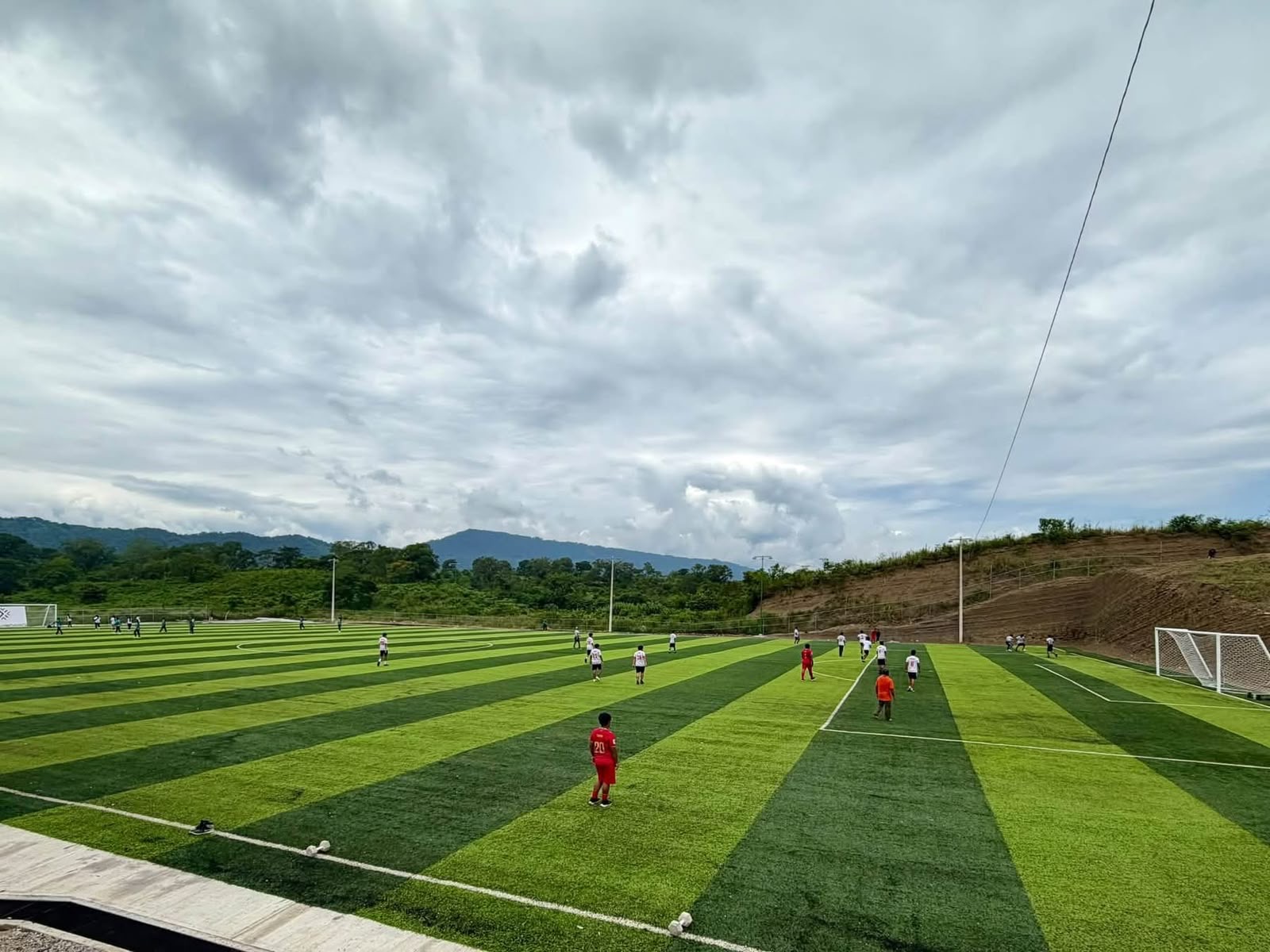 Cancha de fútbol de césped sintético con jugadores durante un partido y montañas al fondo, en un espacio deportivo de San Luis Potosí.