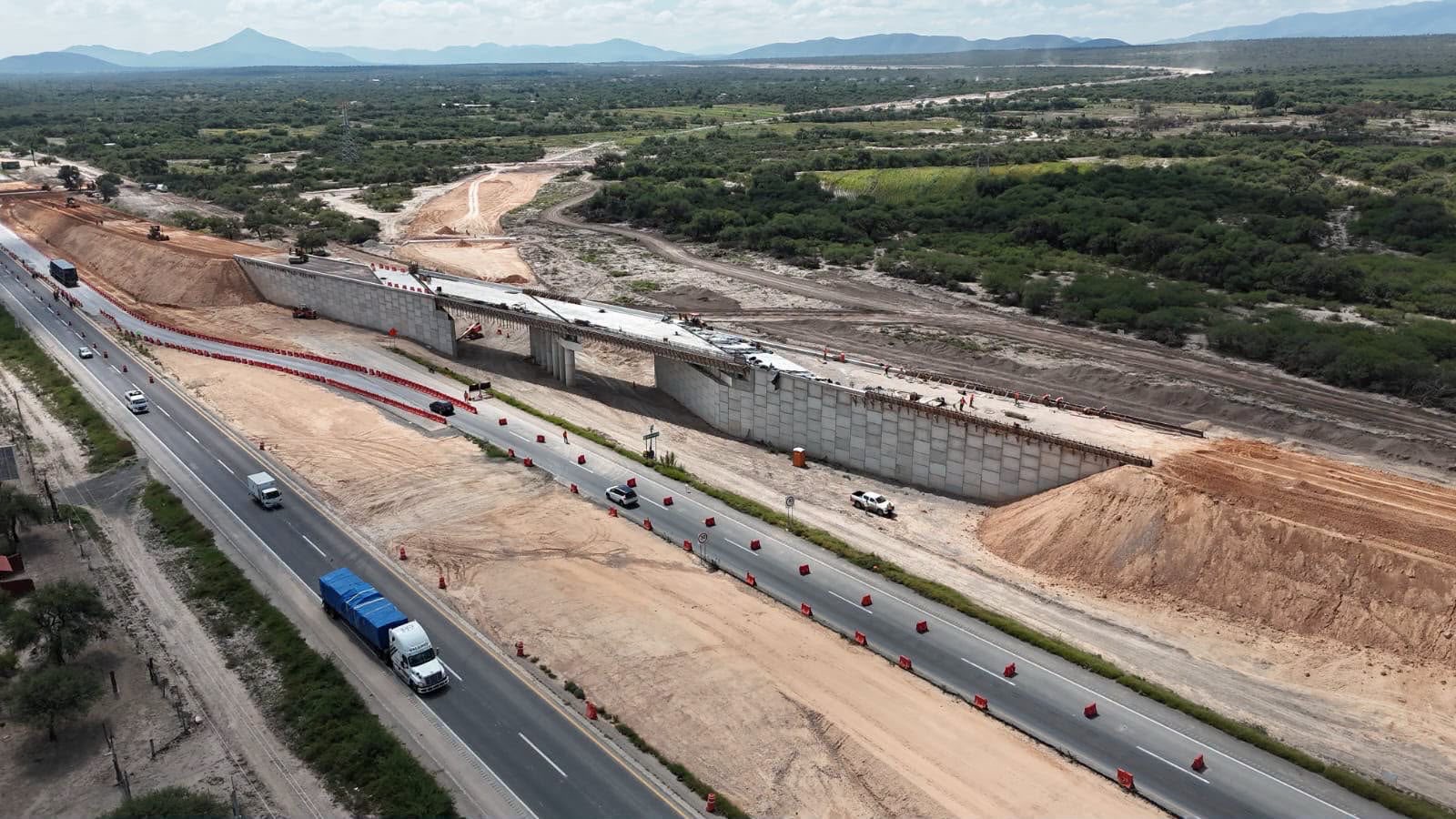 Vista aérea de las obras de construcción de un puente y sus accesos en la autopista San Luis–Matehuala, con circulación vehicular en la carretera contigua.