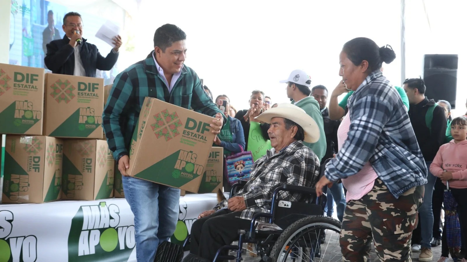 Durante una entrega de apoyos del DIF Estatal, un hombre sostiene una caja de asistencia alimentaria frente a una persona en silla de ruedas acompañada por una mujer.