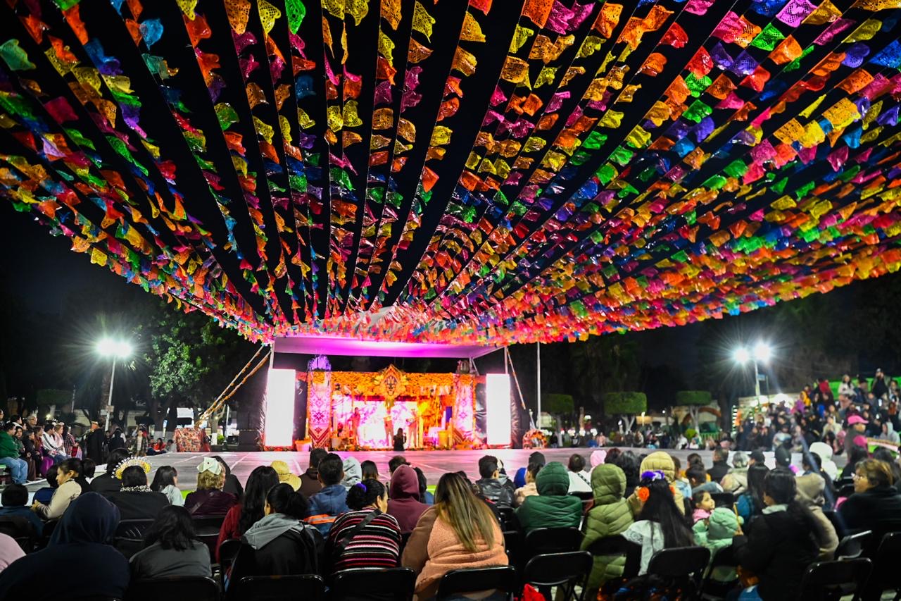 Familias reunidas frente al escenario decorado con papel picado de colores durante las actividades de Xantolo en la Plaza Principal de Soledad de Graciano Sánchez.